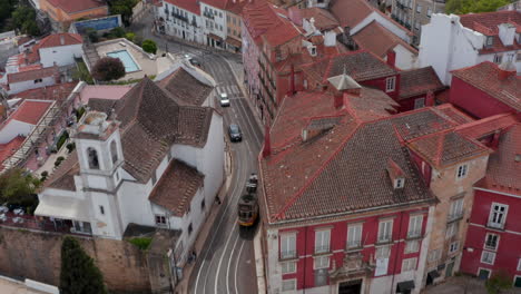 Following-aerial-shot-of-colorful-yellow-traditional-tram-driving-on-the-tracks-between-houses-in-city-center-of-Lisbon,-Portugal