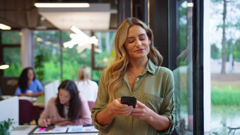 Mature-Businesswoman-Standing-By-Window-In-Busy-Office-Texting-Or-Browsing-On-Mobile-Phone