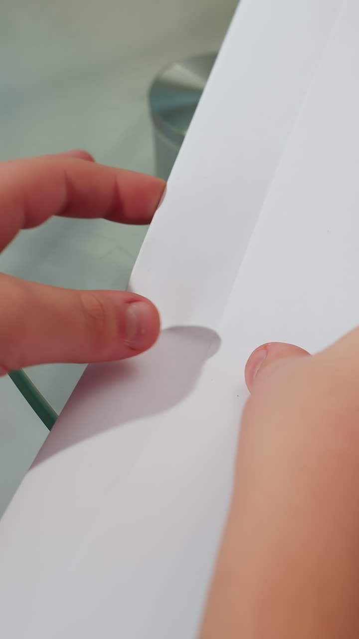 Primer plano de un niño blanco doblando una tira de papel sobre una mesa de cristal; se ven sus pequeñas manos presionando con cuidado el pliegue y el borde texturizado; luz de día tranquila; momento de tranquilidad creando con precisión y paciencia