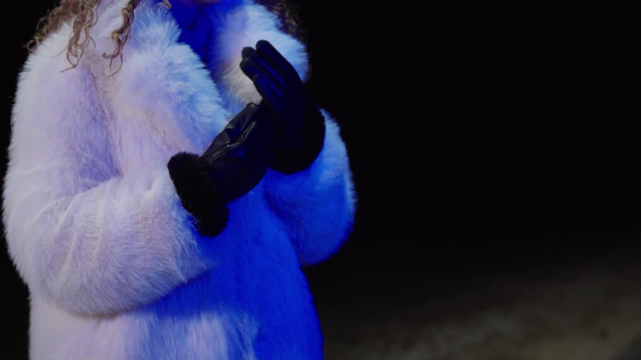 Primo piano di una donna che indossa un cappotto di pelliccia bianco mentre si infila guanti di pelle neri sotto una luce notturna blu. Concept per moda invernale, stile lussuoso, preparazione e estetica della vita notturna.