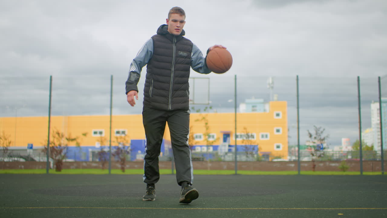 Giovane uomo palleggia con un pallone da basket all'aperto su un campo urbano recintato con sfondo industriale giallo e cielo nuvoloso, indossa un gilet imbottito e scarpe da ginnastica, cammina e fa rimbalzare la palla durante un allenamento individuale, concentrato