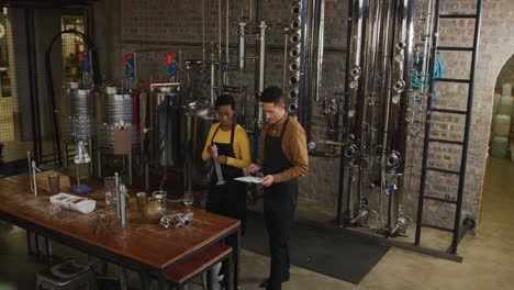 diverse male and female colleague at gin distillery inspecting equipment, talking and making notes