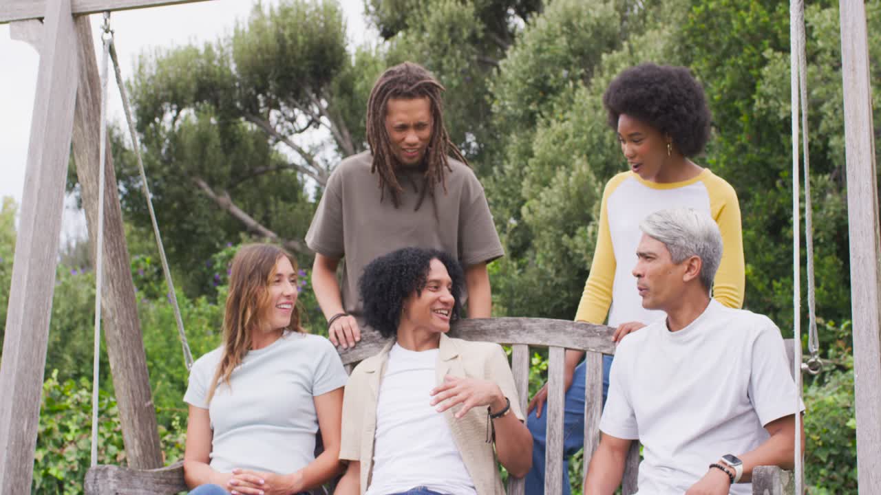 Diverse friends sitting on wooden swing bench in garden, mature man leaning back sharing story