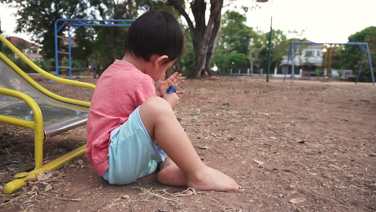 East Asian boy kid sitting on the floor of an outdoor playground by the slider trying to clean his hands from dust and soil