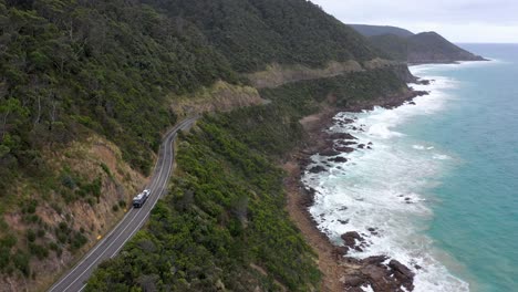 Car-with-caravan-drives-on-Great-Ocean-Road-next-to-coastal-cliffs,-Victoria,-Australia