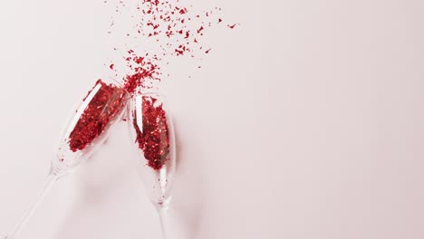 Video-of-two-champagne-glasses-spilling-red-glitter-confetti,-on-white-background-with-copy-space