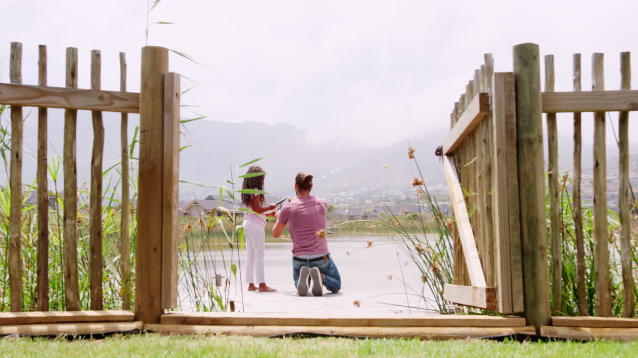 Rear View Of Father And Daughter Fishing In Lake From Wooden Jetty