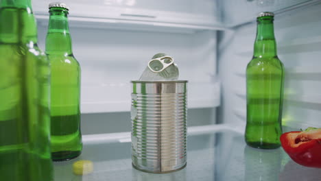 young man looking inside refrigerator empty except for open tin can and bottles of beer on shelf