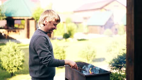joven de pie cerca de la casa de verano prepara los carbones en la barbacoa para cocinar pilaf