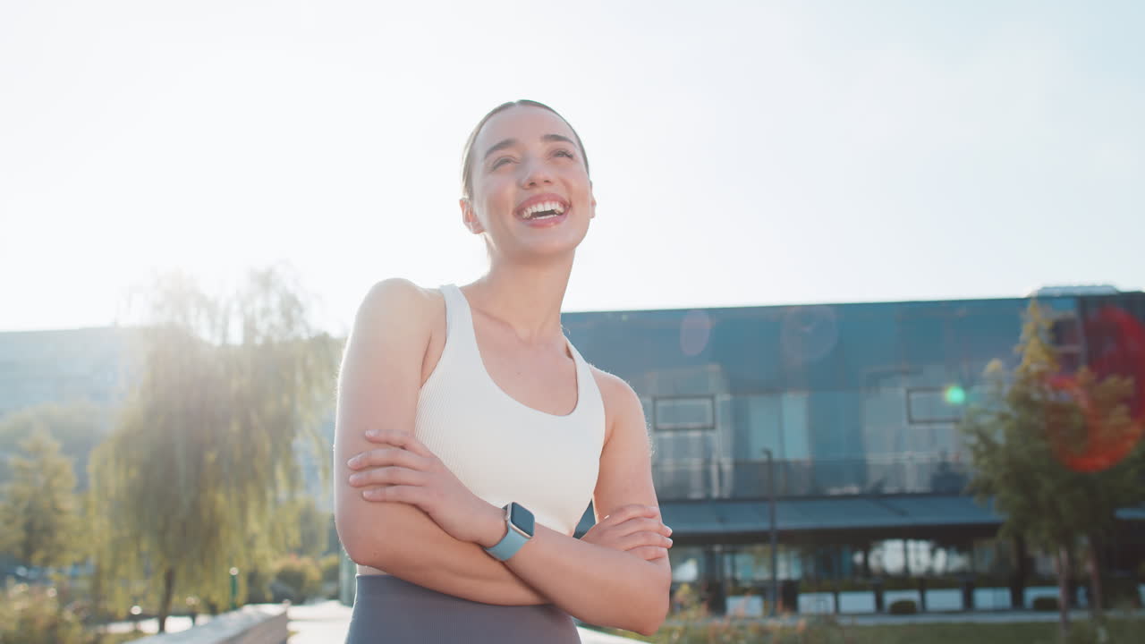 Portrait of laughing caucasian young sporty woman in activewear standing arms crossed in city park