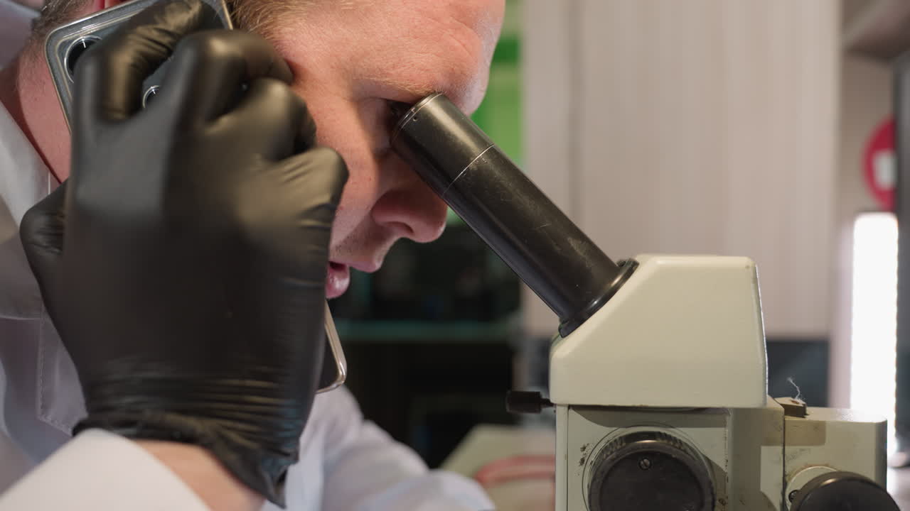 A close-up head view of a technician wearing black gloves and a white lab coat, observing through a microscope while talking on the phone