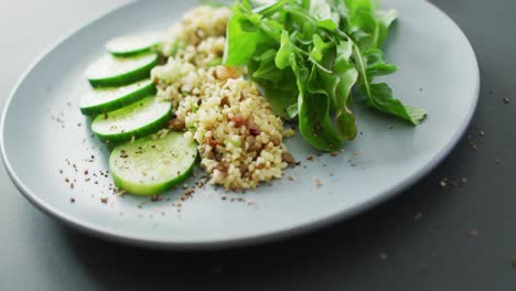 Video-of-fresh-sliced-cucumber,-leaves-and-grains-salad-on-grey-plate-over-dark-grey-background