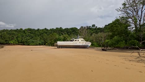 Camera moves laterally past a beached, abandoned boat on a wide sandy shore with tropical trees under bright daylight in Ko Phayam, Thailand