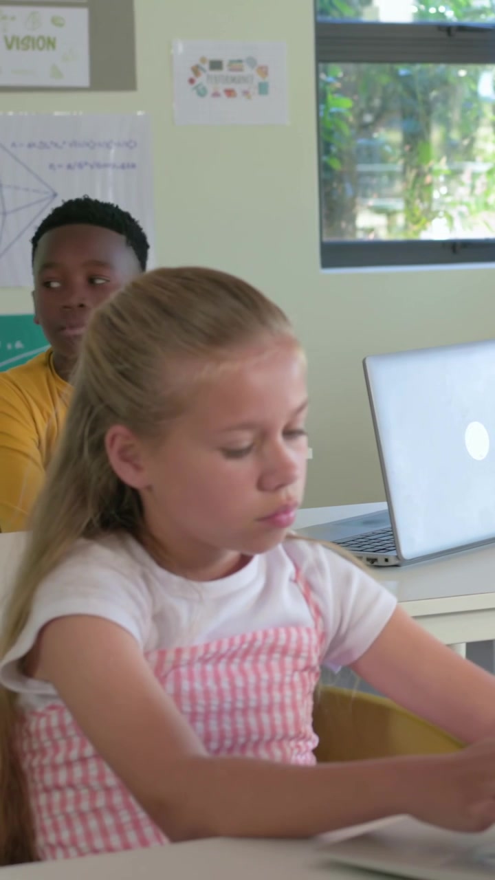 Video verticale: La telecamera si sposta su una ragazza in grembiule rosa seduta a scuola che lavora su documenti con un laptop