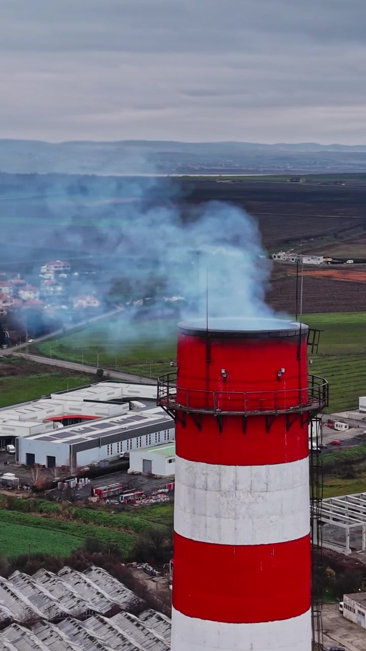 Il fumo si alza da un'alta ciminiera in una zona industriale vicino a campi ed edifici