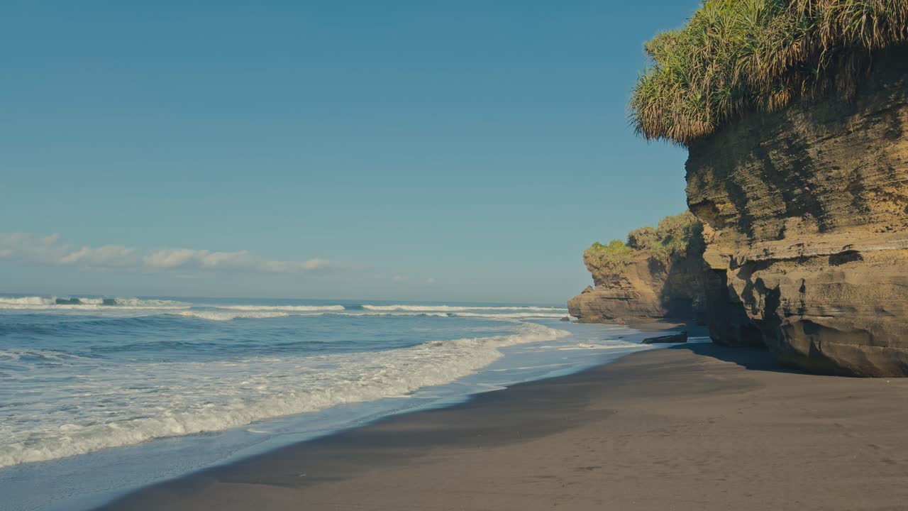 Pemandangan damai pantai pasir hitam dengan tebing pantai kapur dan ombak tenang bergulir di hari tropis yang cerah dan tenang dengan langit biru jernih, menciptakan lanskap tepi laut yang tenang dan indah
