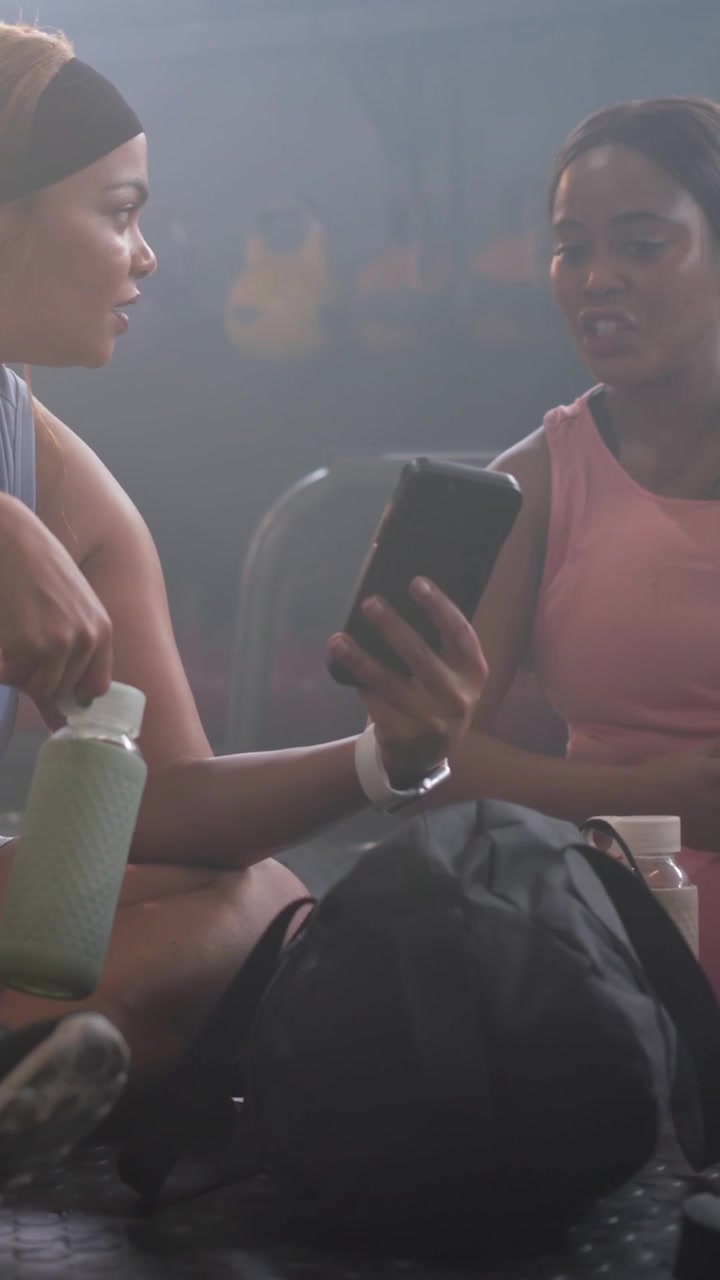 Vertical video: Post-workout woman in blue set handing phone at gym, holding green bottle and towel