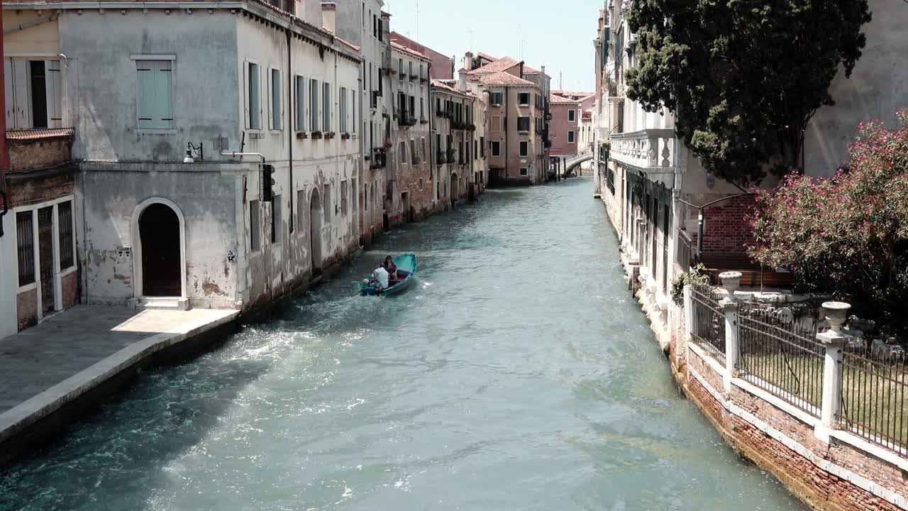 Veduta diurna lungo uno stretto canale a Venezia, Italia, con un piccolo motoscafo che si muove nell'acqua turchese tra vecchi edifici residenziali e passerelle sul lungomare.