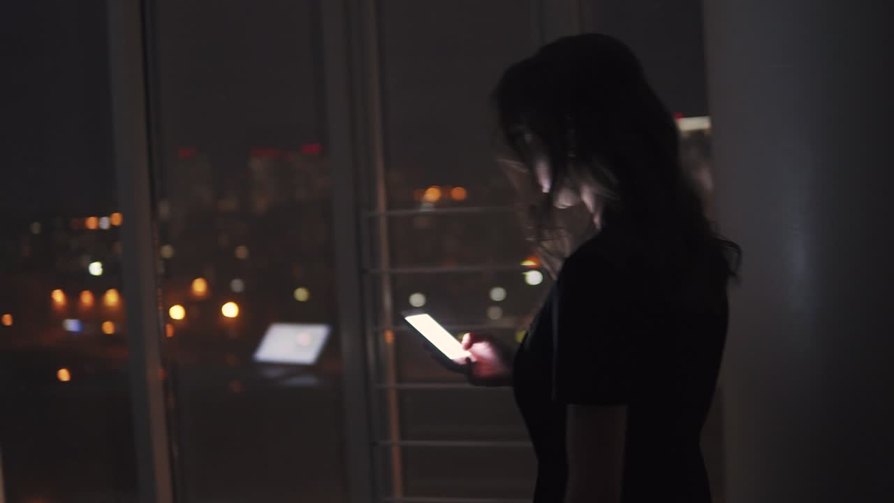 donna d'affari che cammina in un ufficio buio con uno smartphone in mano. ritratto di una ragazza sul posto di lavoro sullo sfondo della città notturna.