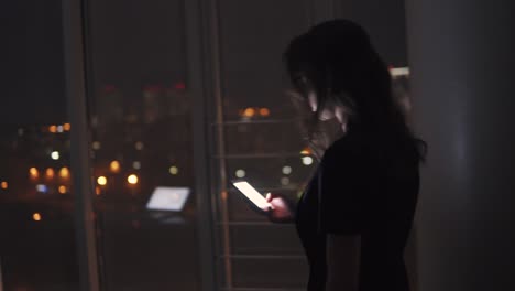 businesswoman walking in a dark office with a smartphone in her hands. portrait of a girl at the workplace on the background of the night city.