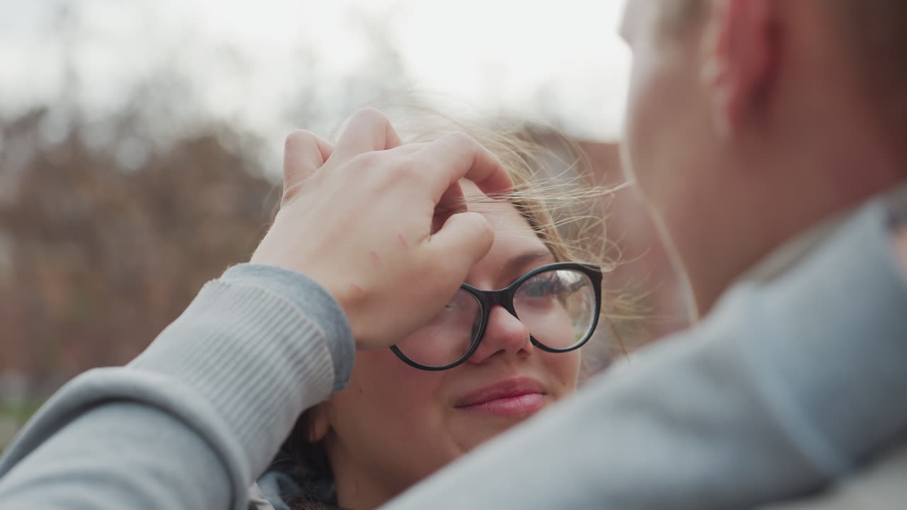 Close up of young couple gazing at each other as man gently adjusts woman hair fluttering in wind, sharing tender romantic moment with soft smiles and deep connection outdoors on breezy day