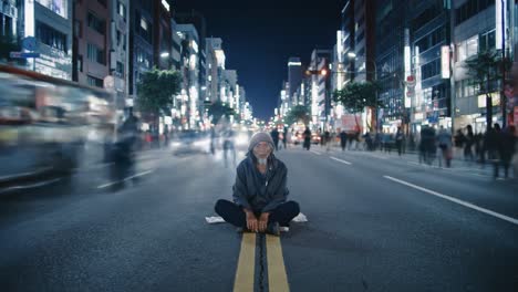 a man sits cross-legged on the road in a bustling city at night. bright lights from buildings and passing vehicles create a lively atmosphere, highlighting his solitude in the crowd.