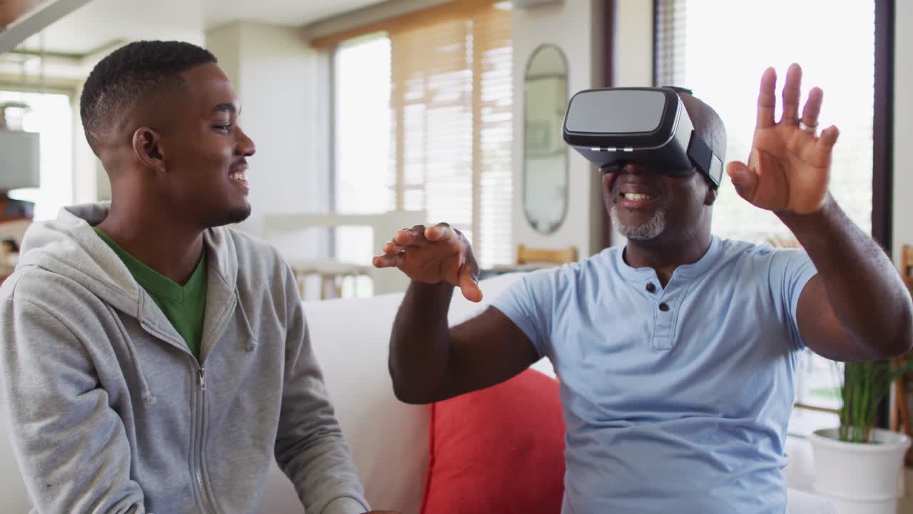 African american senior father sitting on a couch using a vr headset with his teenage son smiling