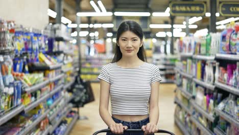 Happy-Asian-girl-walks-around-the-supermarket-with-a-shopping-cart-and-looking-at-the-camera.-Shopping-trip.-daily-business