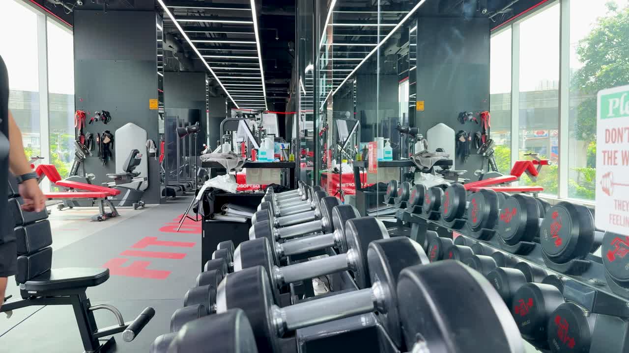 Two men in a gym select dumbbells for a bicep workout. Bright lighting and modern equipment create an energetic atmosphere