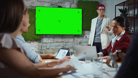 reunión en la sala de conferencias de la oficina: la estratega jefe de la compañía hace una presentación de televisión en pantalla verde a un equipo diverso de empresarios multiétnicos. usando el monitor de televisión de pared de chroma key