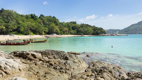 a tranquil beach in phuket with clear turquoise waters, rocky foreground, and lush greenery under bright daylight