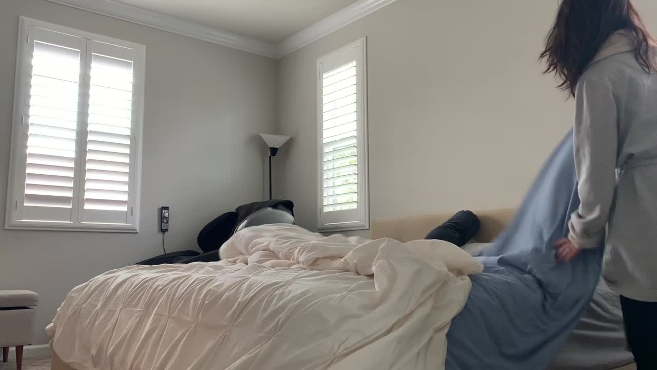 Woman getting ready for a brand new start to a day. She stretches, gets up and out of bed, and fixes her bed coverings. White simple room with natural sunlight shining through window blinds.