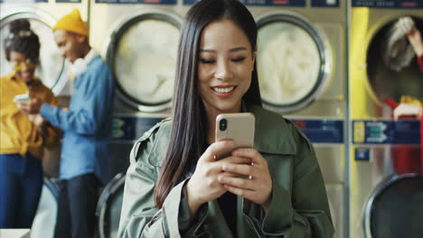 young pretty and stylish girl in yellow glasses standing in laundry service room and tapping on smartphone 1