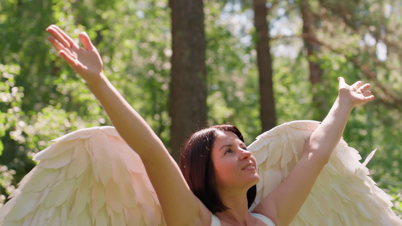 Magical creature with soft white angel wings lifts hands gracefully toward shining sunlight in lush green forest, surrounded by glowing leaves and golden warmth