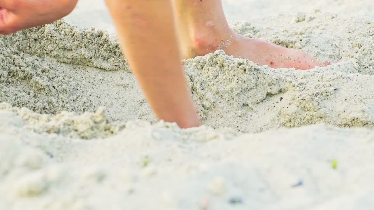A kid gets up in a sandy pit with his legs to measure it and continues to expand its length and width. Close-up.