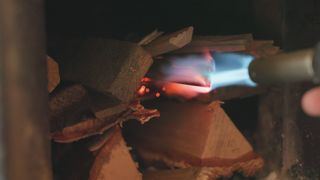 Close up inside dark chimney shows person using igniter flame to light stacked dry fire wood, bright spark igniting bark edges, beginning process of kindling fire for indoor warmth