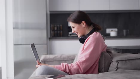 Woman-At-Home-Sitting-At-Kitchen-Sofa-Making-Video-Call-On-Laptop-Wearing-Headphones