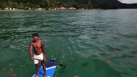 Aerial-drone-pivot-circular-bird's-eye-view-of-man-exercising-on-a-sup-paddle-board-in-turquoise-tropical-clear-waters,-with-beach-jungle-and-coastline-in-Thailand