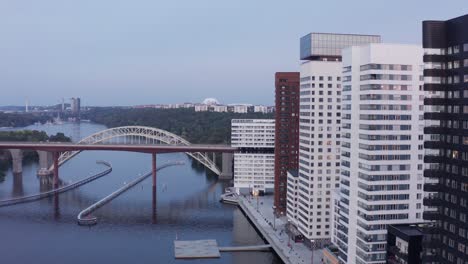 Drone footage high-rise residential buildings with modern design and architecture in &Aring;rstadal, Stockholm with train going by on bridge in the background by the water with trees