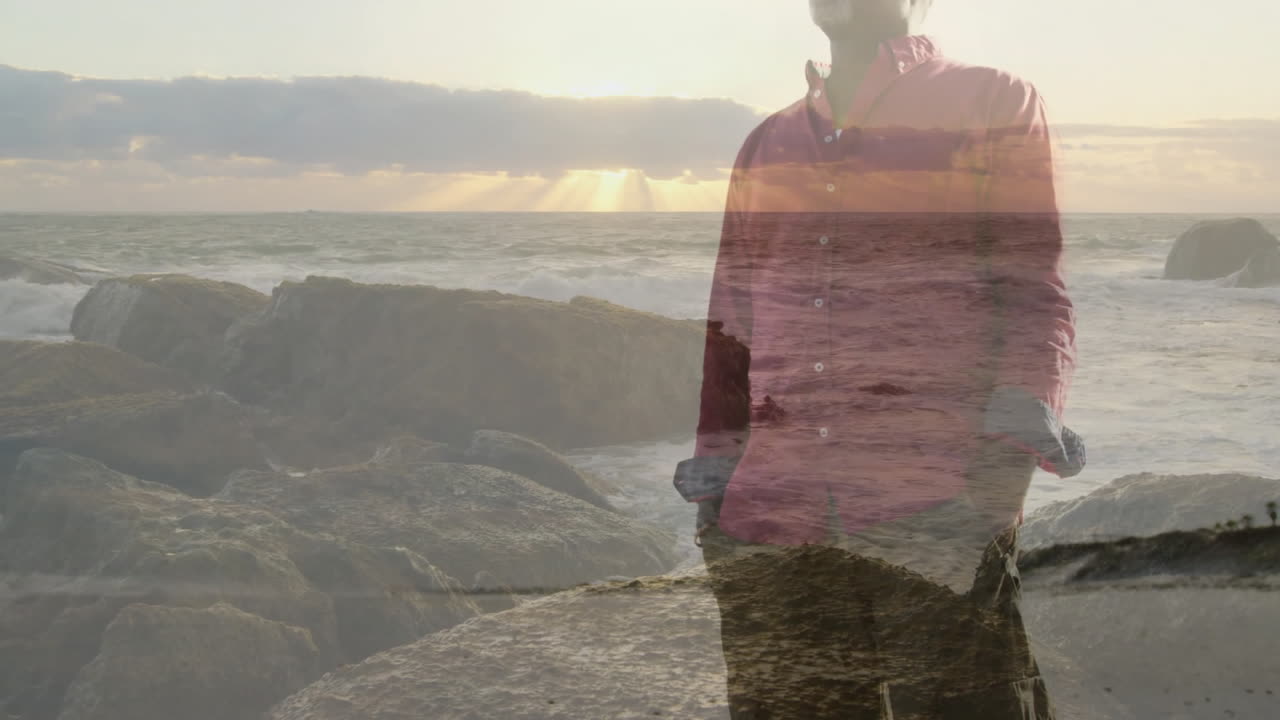 Senior man standing on rocky shoreline at sunset blending with floating charts for finance