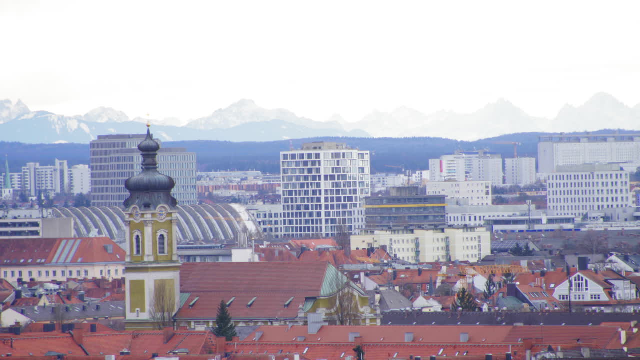 A stunning view of Munich, Germany, featuring a historic church tower in the foreground, modern buildings, and the snow-covered Alps in the background.