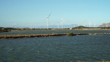 static low pov of wind turbines against blue sky, river in foreground, vietnam