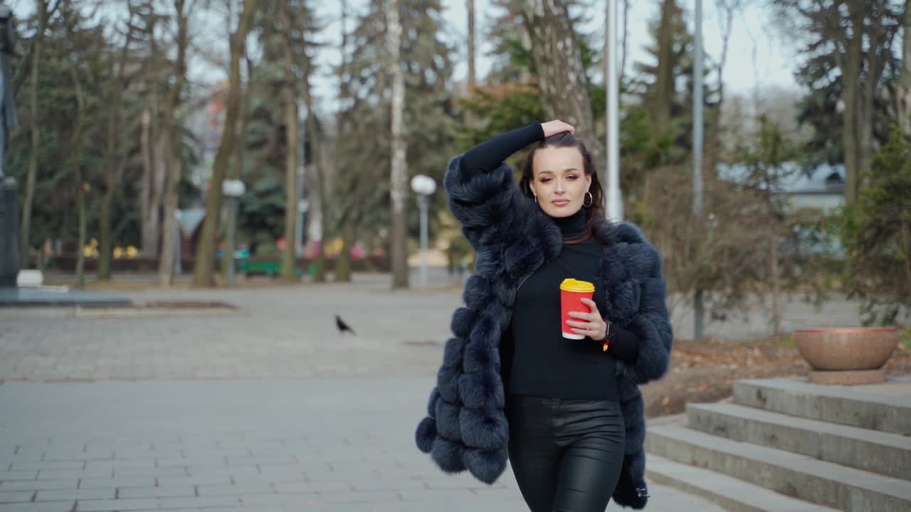 Beautiful young girl in luxury fur coat in the city park. Amazing view of gorgeous lady with a cup of coffee looking at camera outdoors. Slow motion.