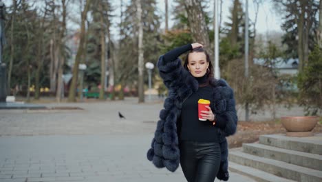 beautiful young girl in luxury fur coat in the city park. amazing view of gorgeous lady with a cup of coffee looking at camera outdoors. slow motion.