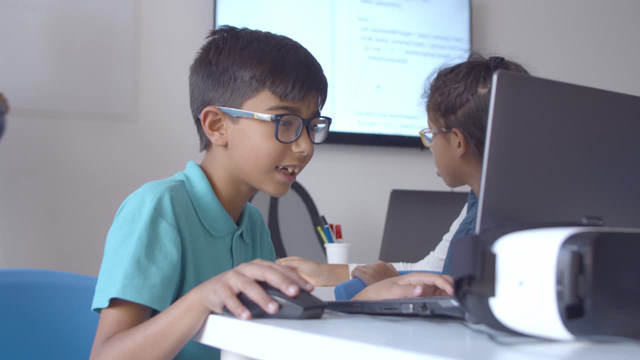 niño alumno emocionado con gafas sentado en escritorios y usando computadora portátil