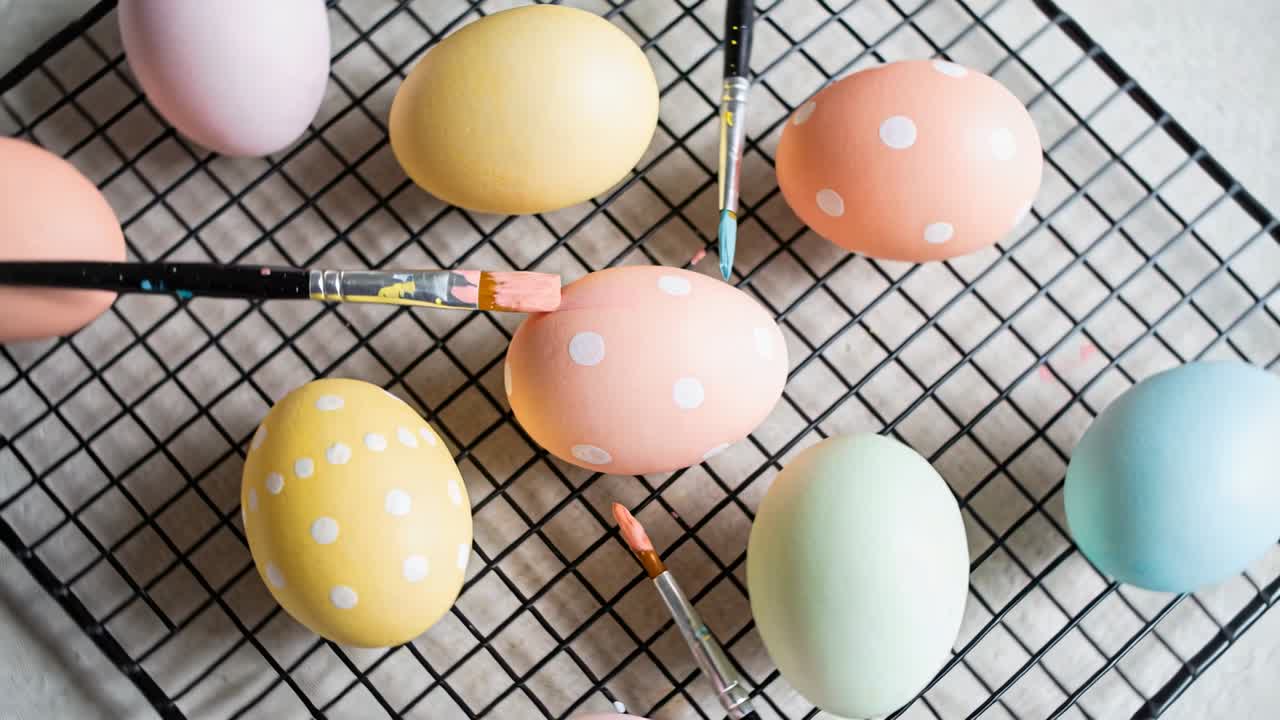 DIY paintbrushes shifting closer, applying white dots on pastel eggs on rack, creating patterns