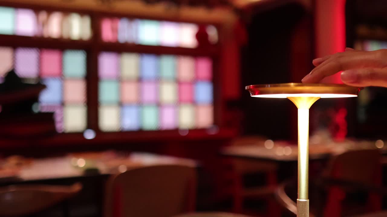 A hand reaches out to activate a sleek, circular table lamp in a warmly lit, modern interior with colorful window panels. Shallow depth of field and smooth camera focus