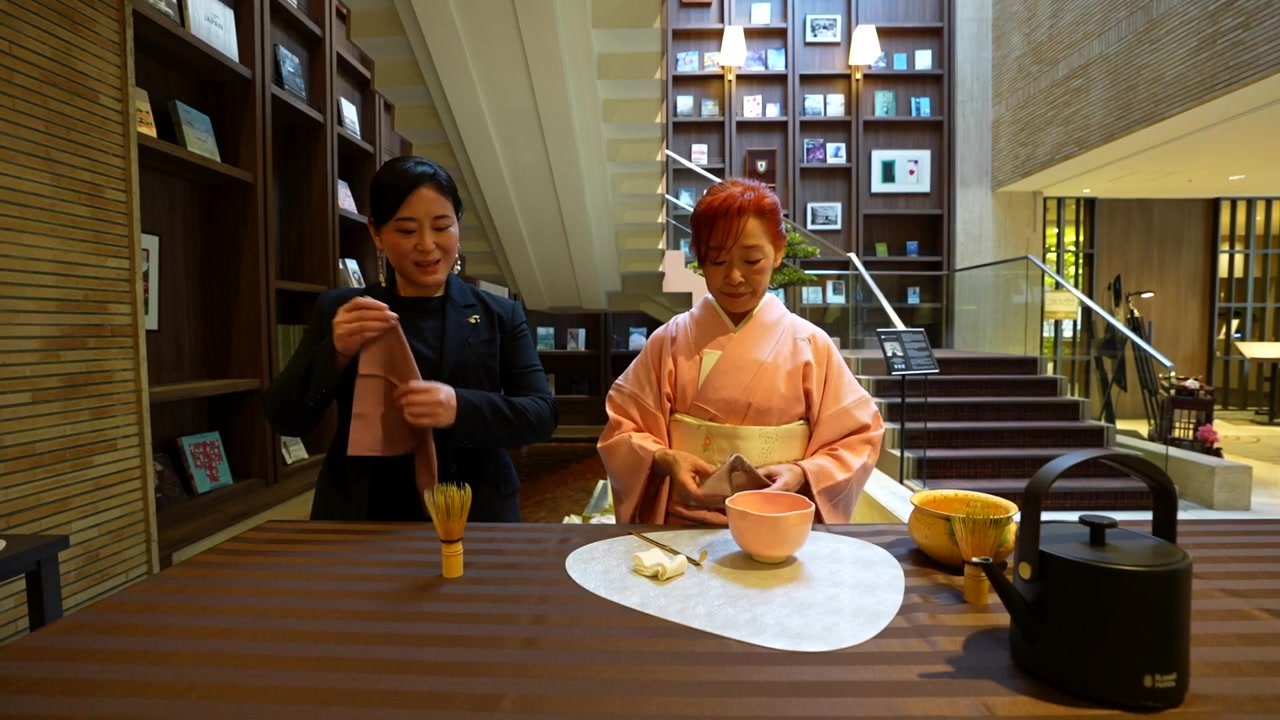 Woman in a traditional pink kimono performing a Japanese tea ceremony with an assistant explaining