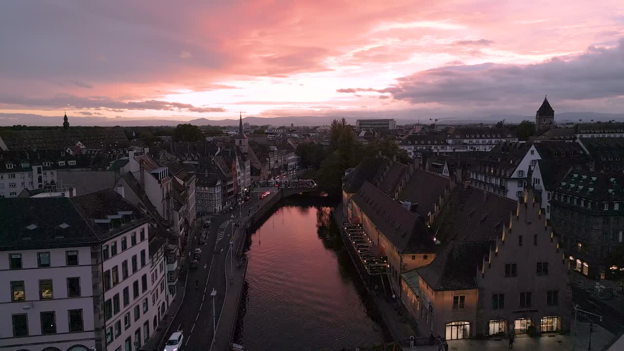 A romantic aerial view of Strasbourg at sunset, with warm pink skies reflecting on the canal waters and historic half-timbered houses lining the serene old town streets