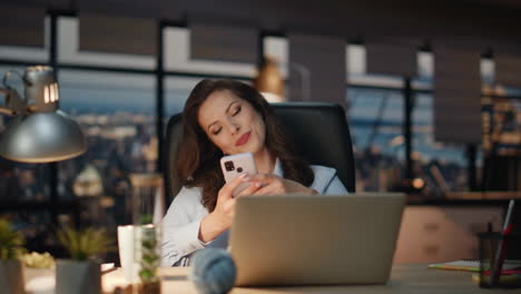 mujer relajada descansando teléfono inteligente noche vista de la ciudad oficina. mujer relajante sillón